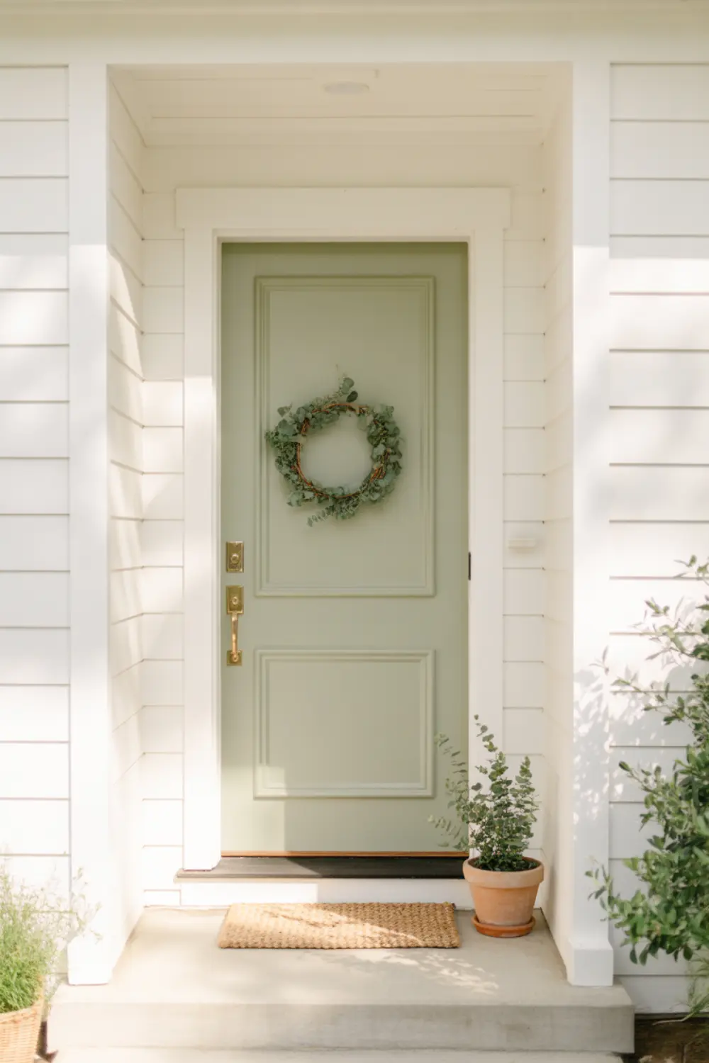Soft sage green front door on a white farmhouse with a cozy welcome mat.